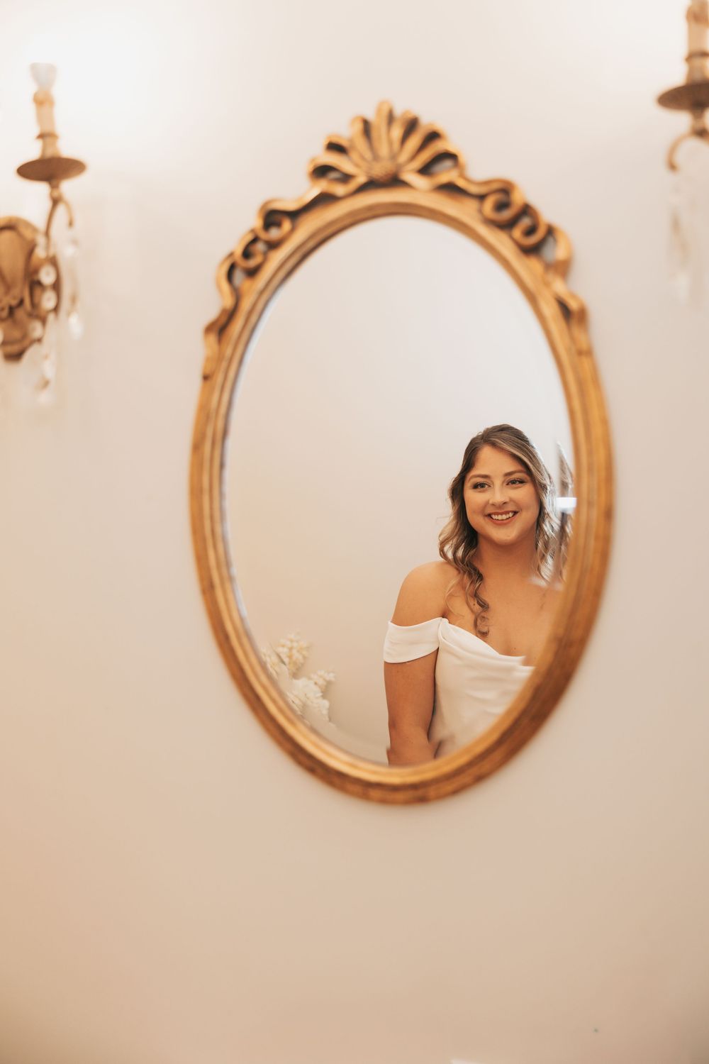 Reflection in an ornate gold oval mirror shows someone wearing an elegant off-shoulder white dress.
