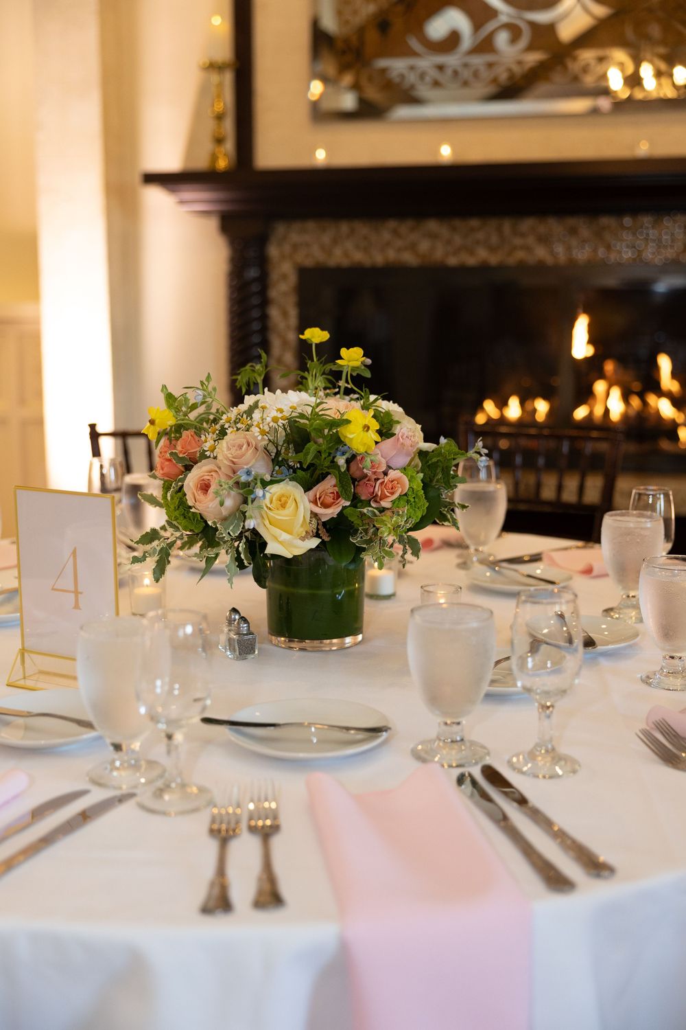 Elegant wedding table setting with pink and yellow floral centerpiece and fireplace in background.