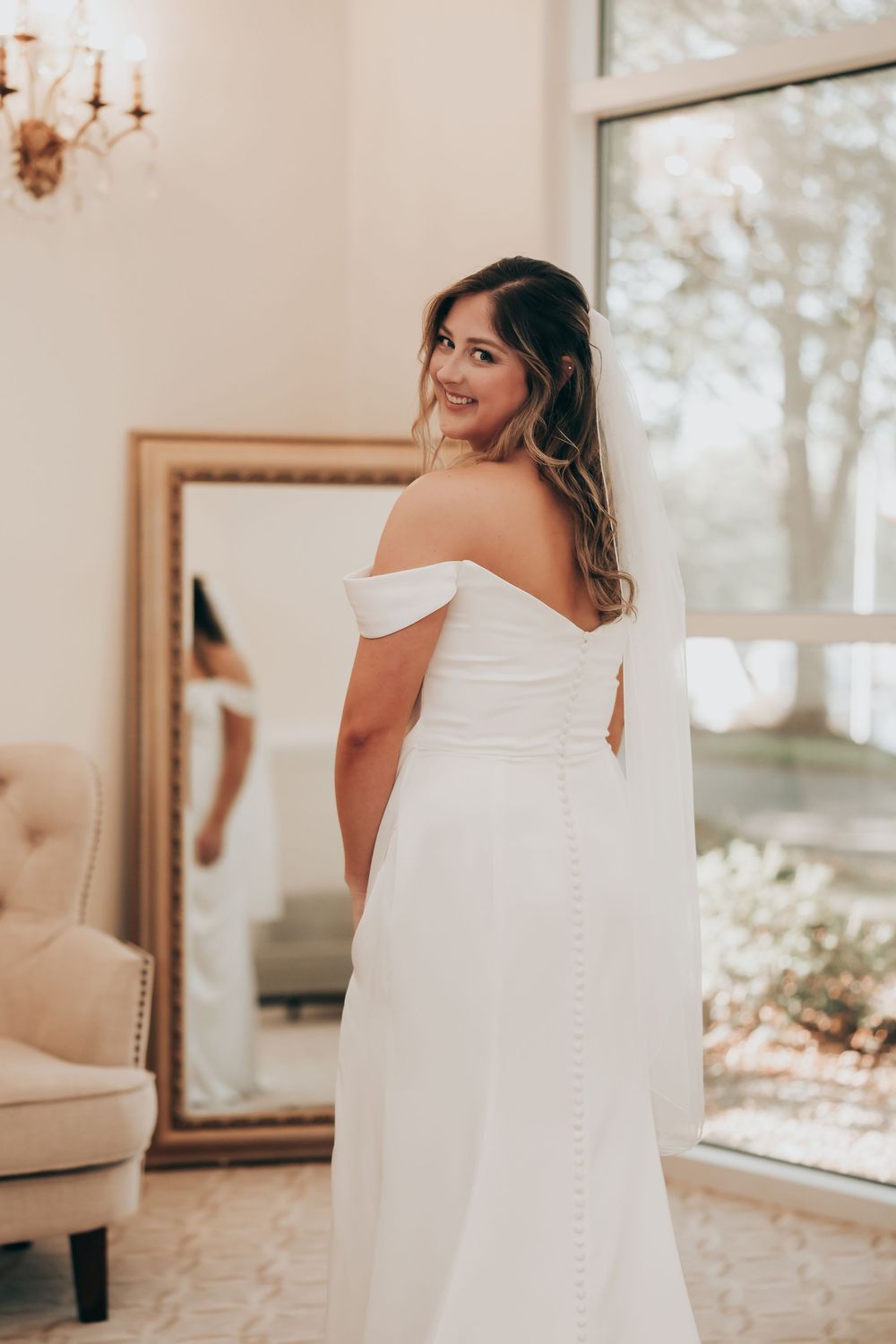 A bride in an elegant off-shoulder white wedding gown looks back over her shoulder in a sunlit room with a large mirror.