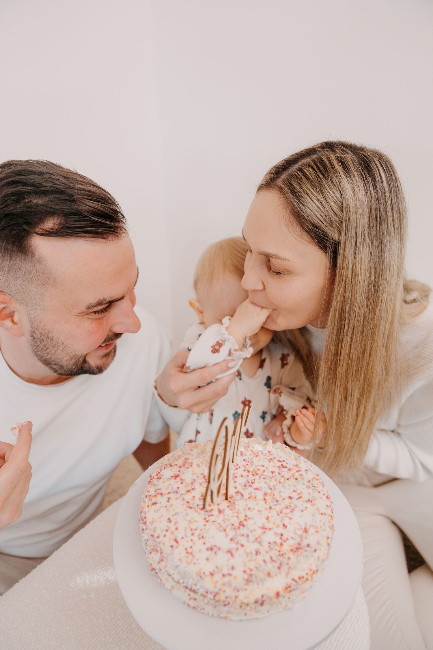 A family celebrates together with a white birthday cake decorated with pink sprinkles and a gold number one topper.