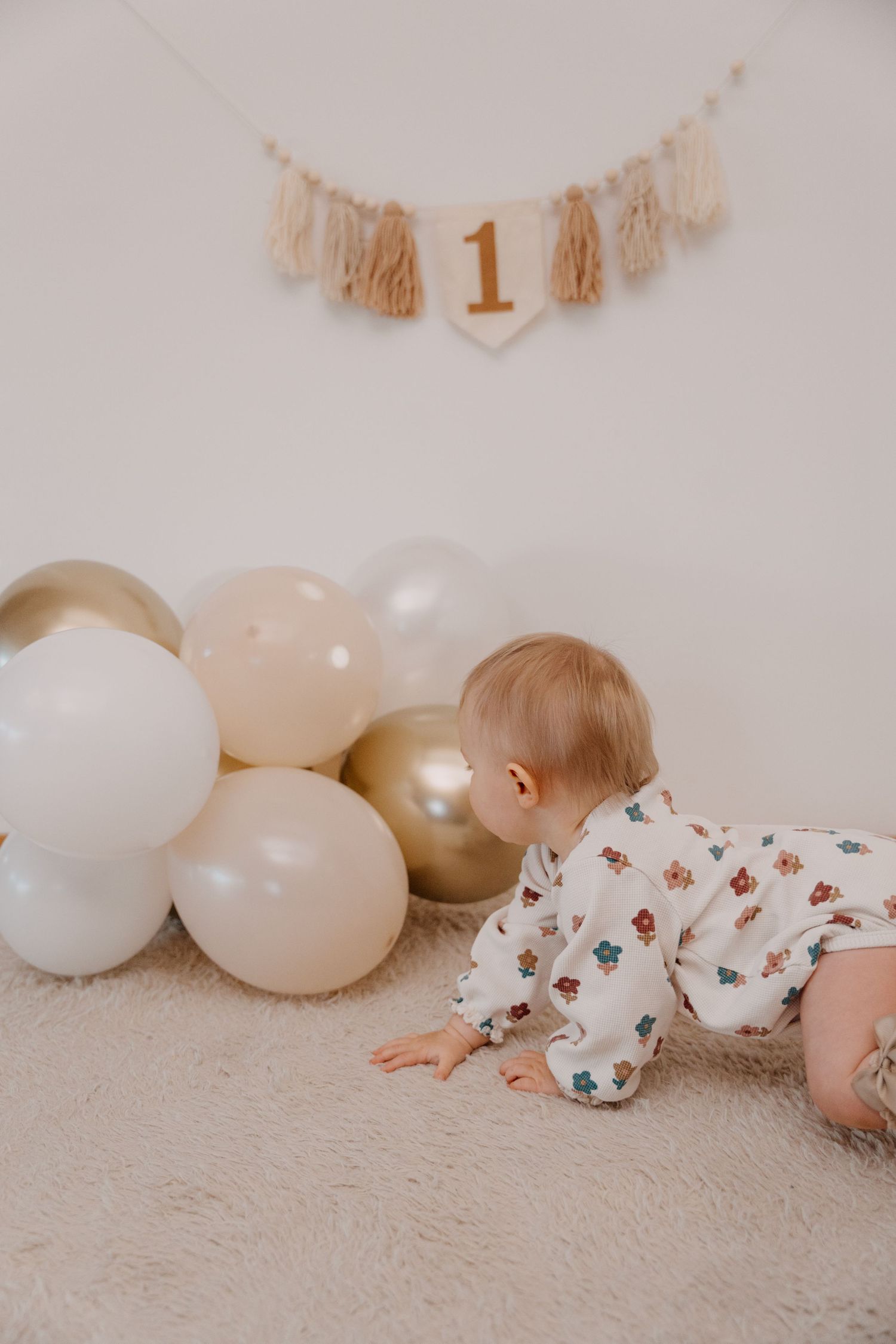Baby in floral onesie crawls on carpet near white balloons and gold number one birthday banner decoration.