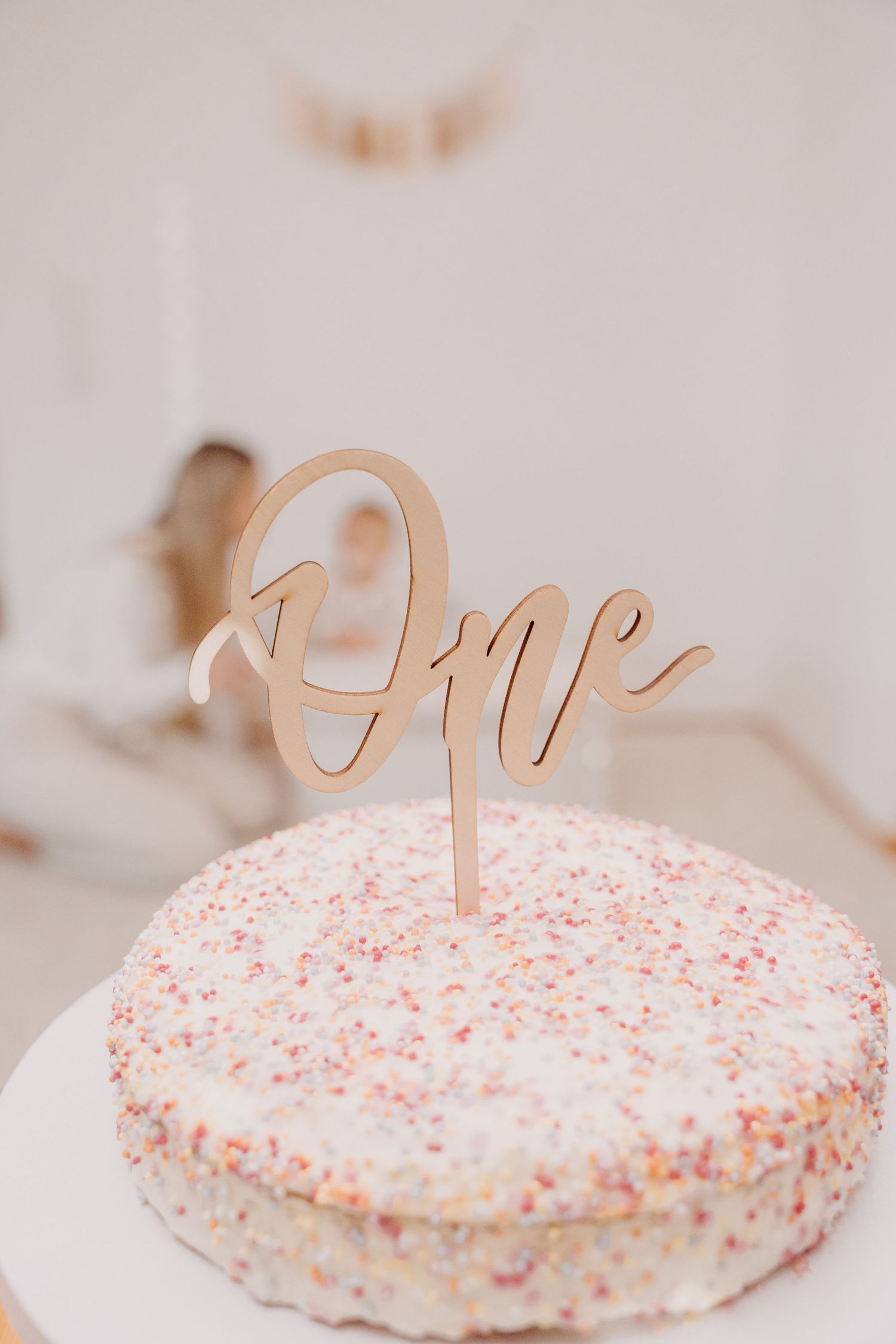 Close-up view of a white frosted cake with pink sprinkles and gold script cake topper decoration.