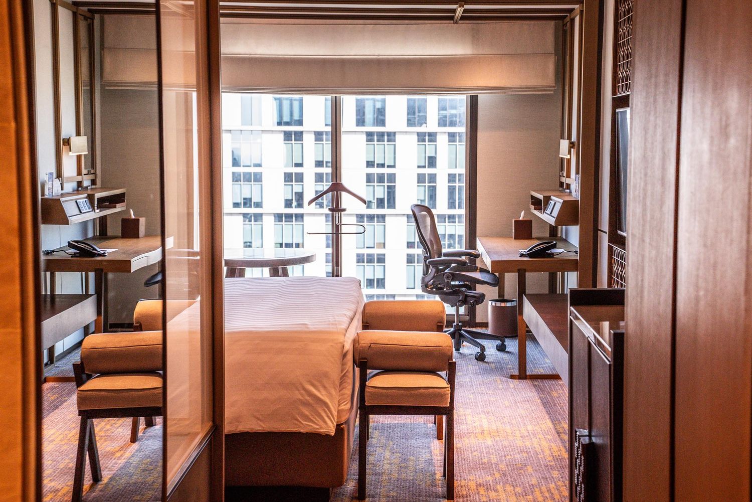 Modern hotel room interior with desk workspace, office chairs, and large windows overlooking city buildings.