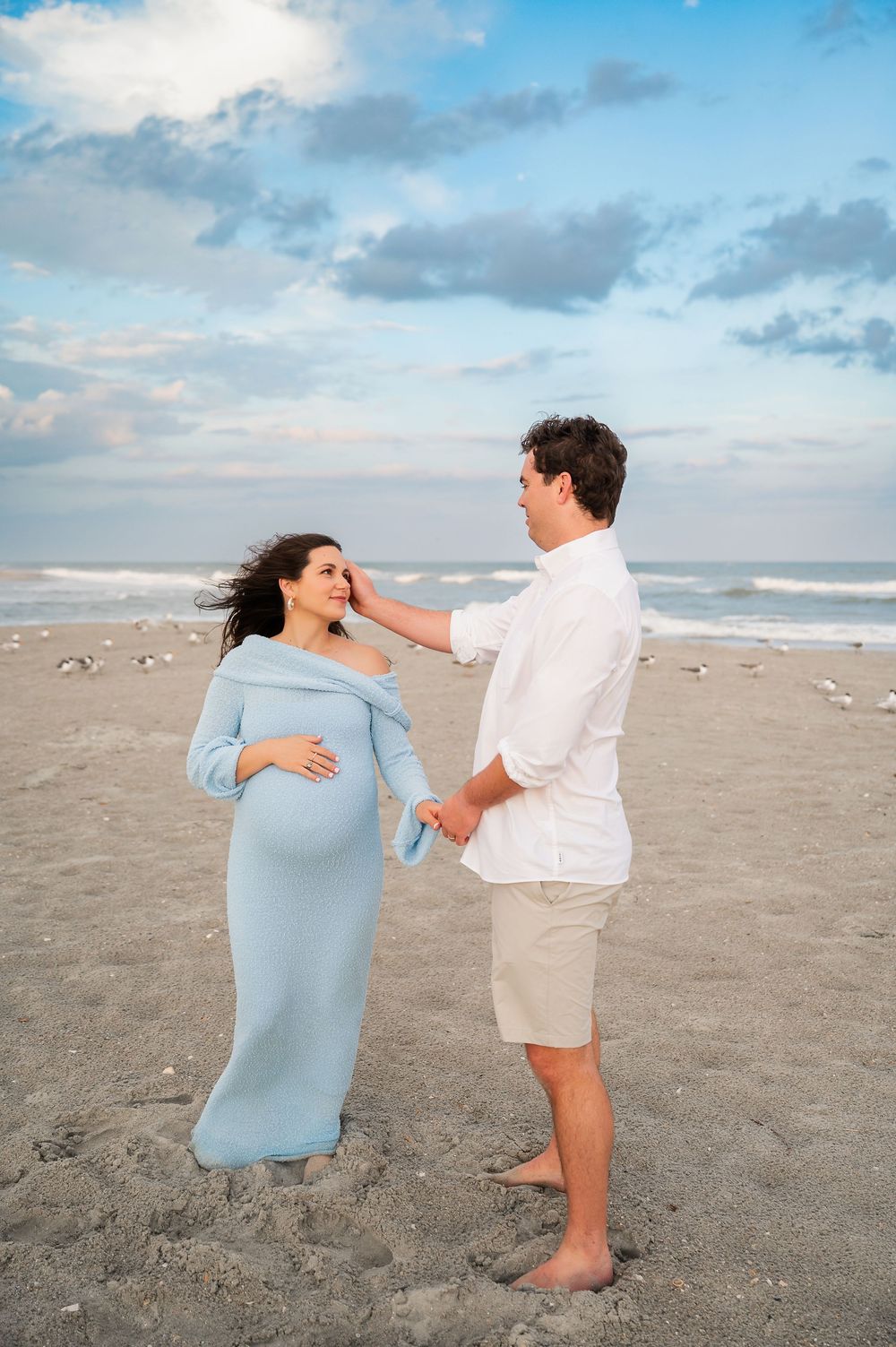 A couple poses playfully on a beach at sunset, with one wearing a light blue dress and the other in white and khaki.