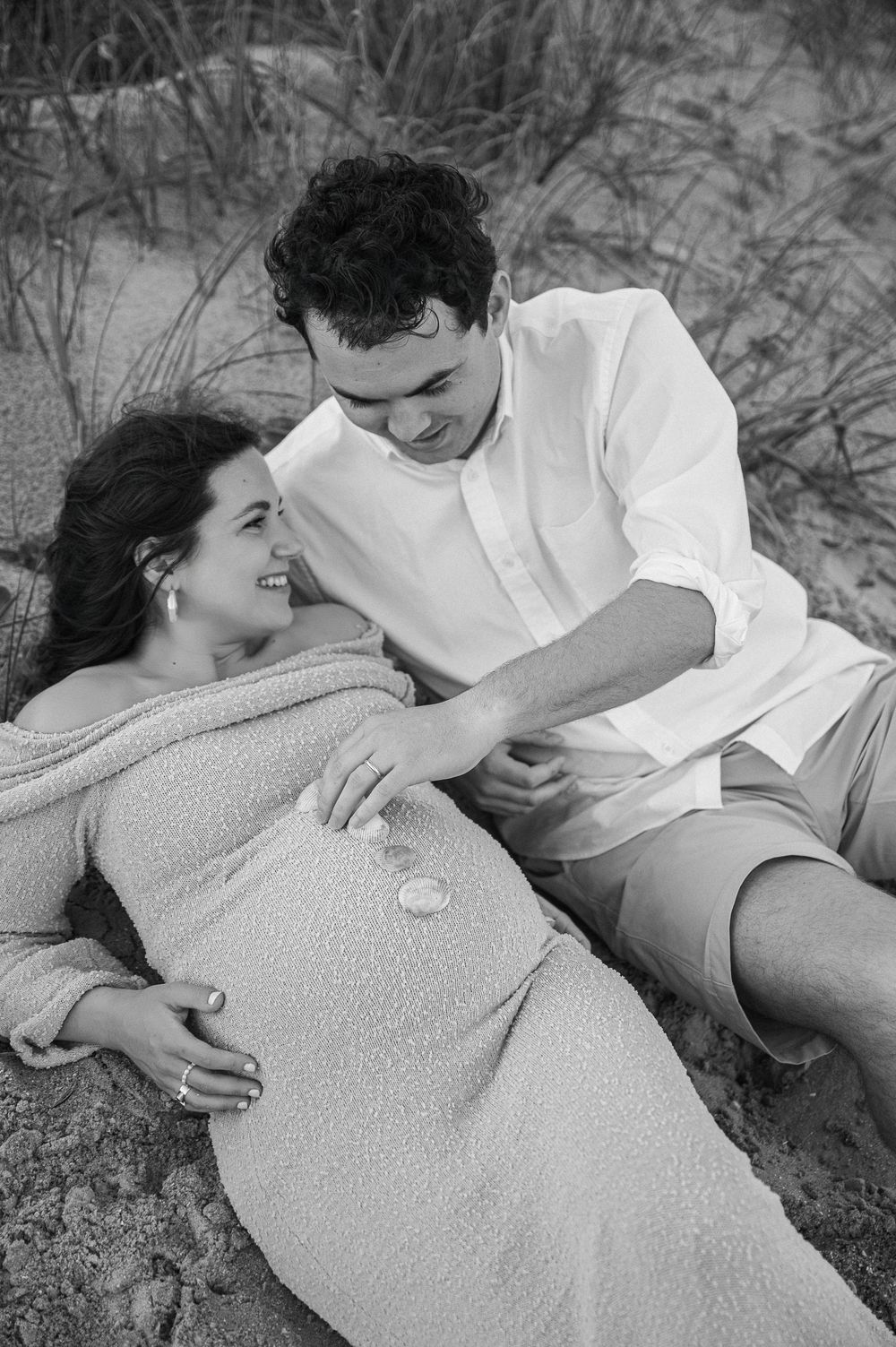 Black and white intimate beach maternity photos showing a couple resting on sandy dunes.