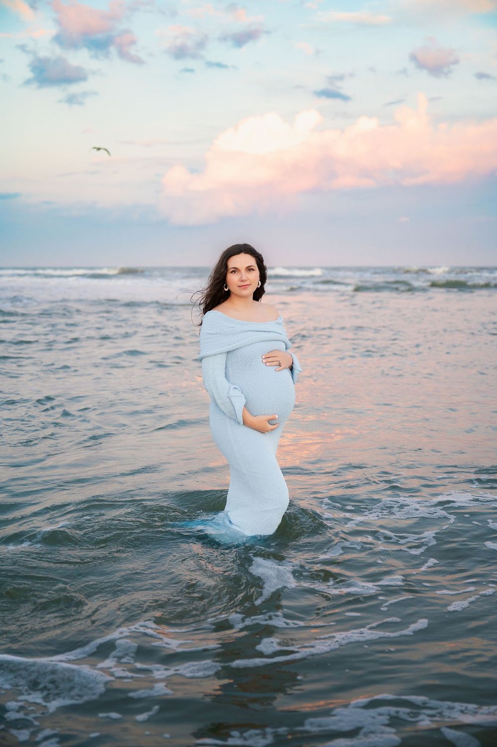 Woman in light blue dress poses in ocean water during sunset for maternity photos.