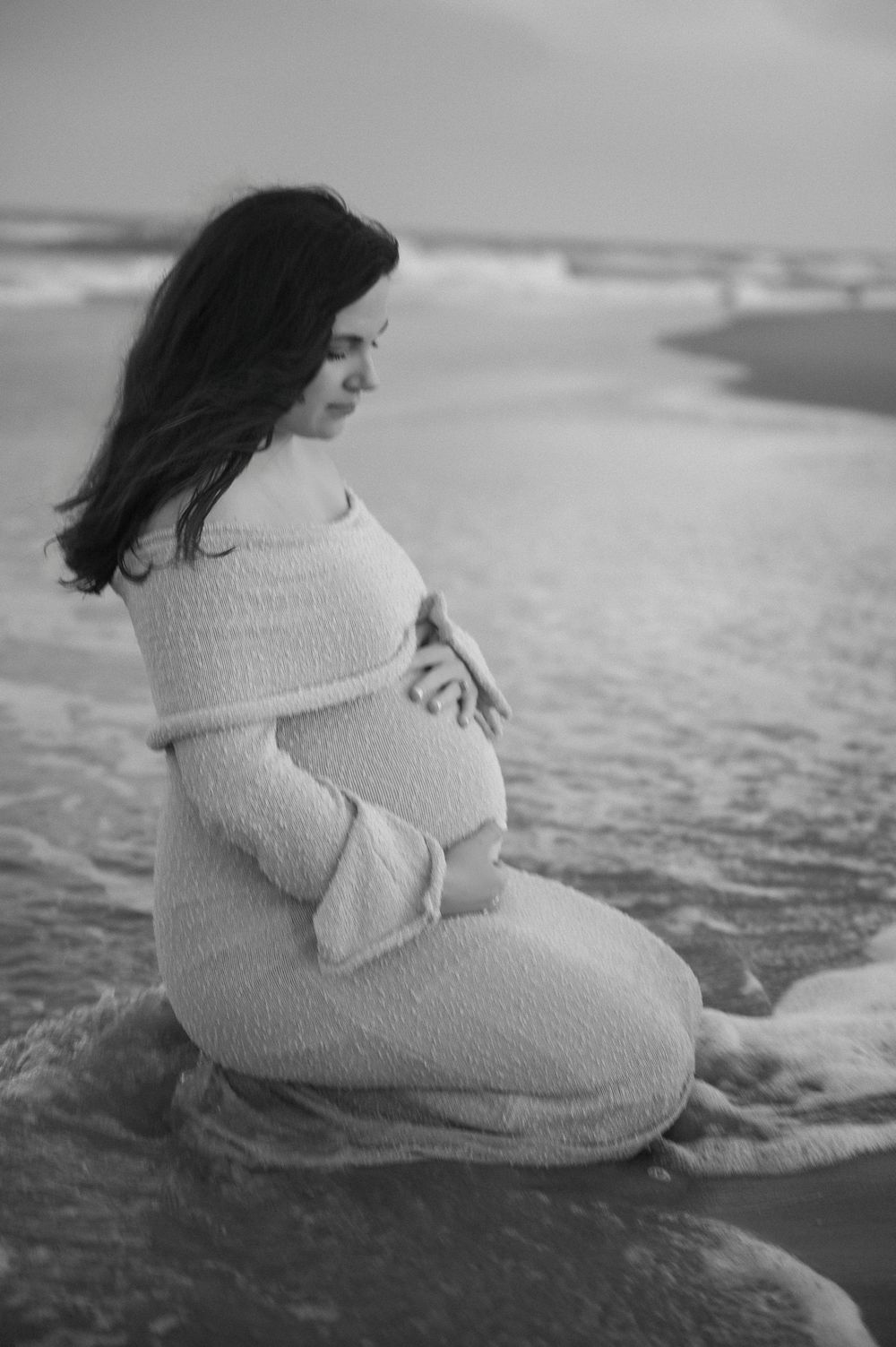 Black and white photo of person in white dress sitting by ocean waves.