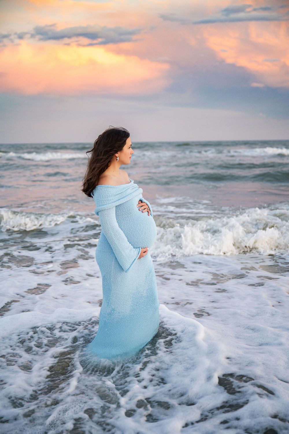Maternity photos at sunset with waves crashing around woman in blue dress on beach.