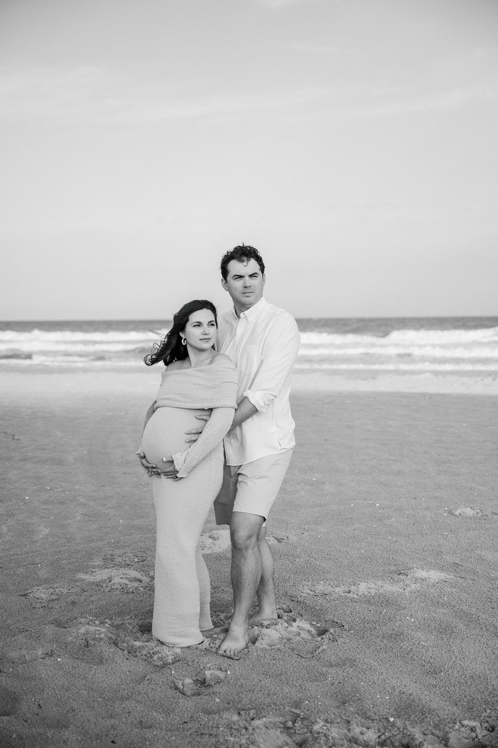 Black and white beach portraits showing couple embracing on sandy shore.