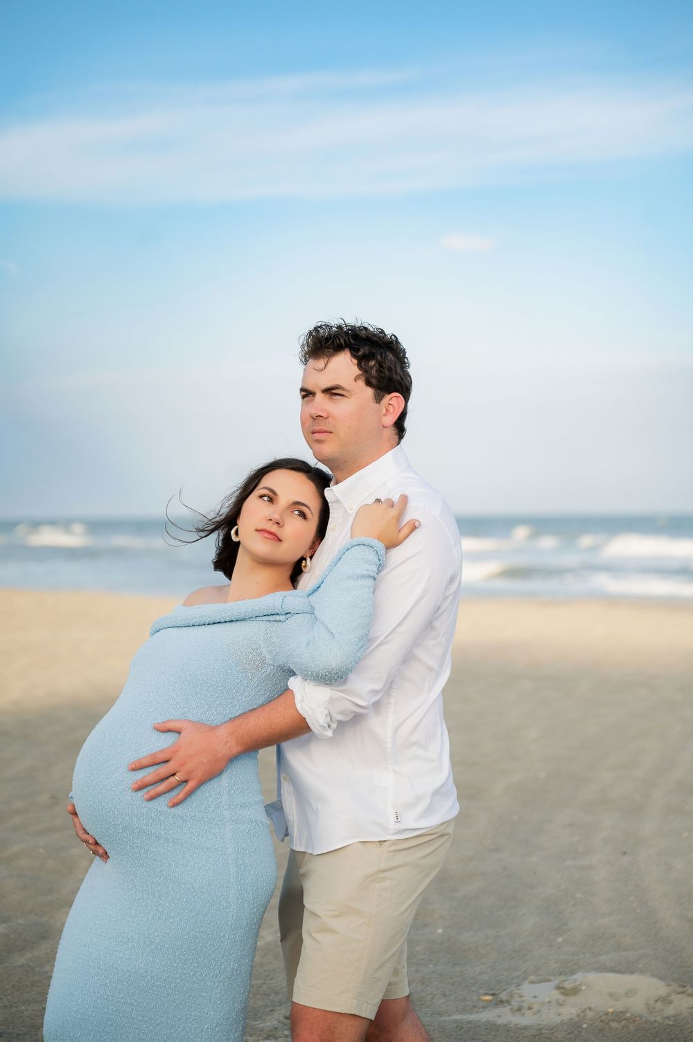 Couple poses together on beach, one wearing light blue dress and other in white shirt and khaki shorts.