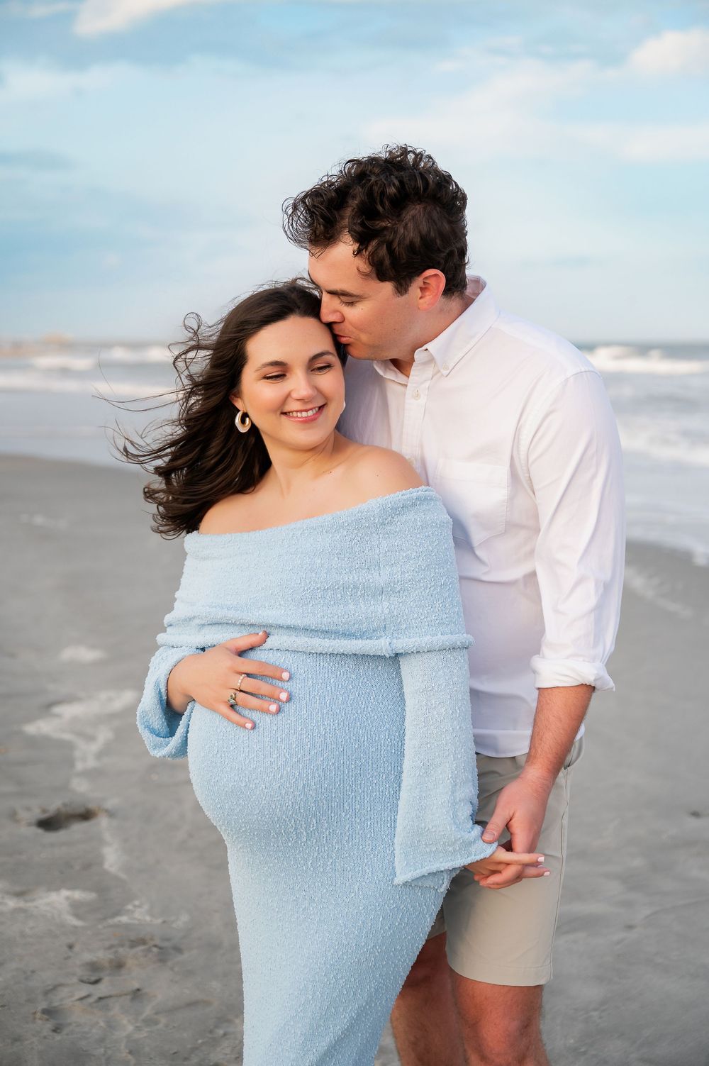 Romantic beach maternity photos with couple in blue dress and white shirt embracing by ocean.