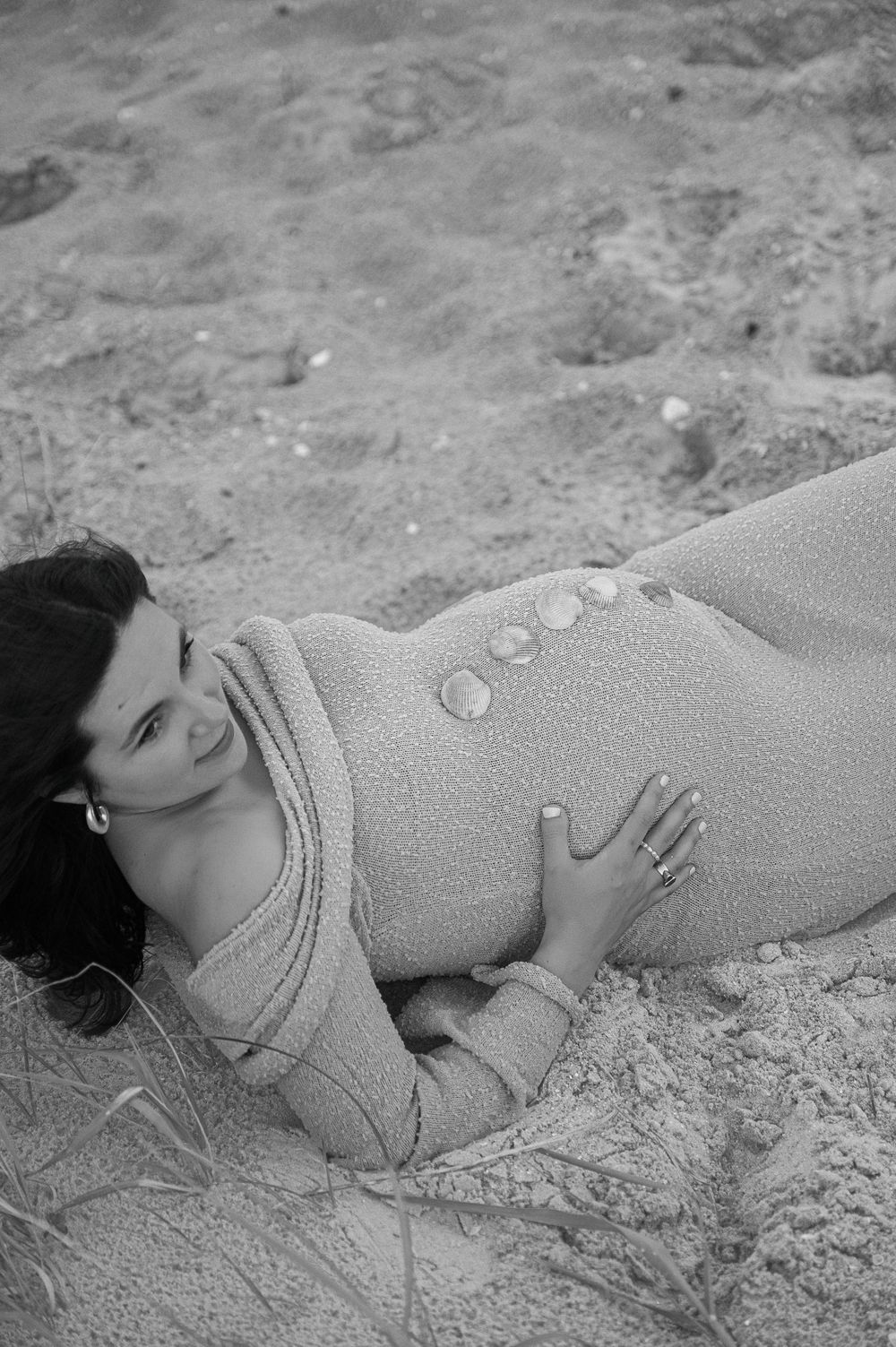 A black and white maternity photo of someone lying in the sand wearing a textured dress.