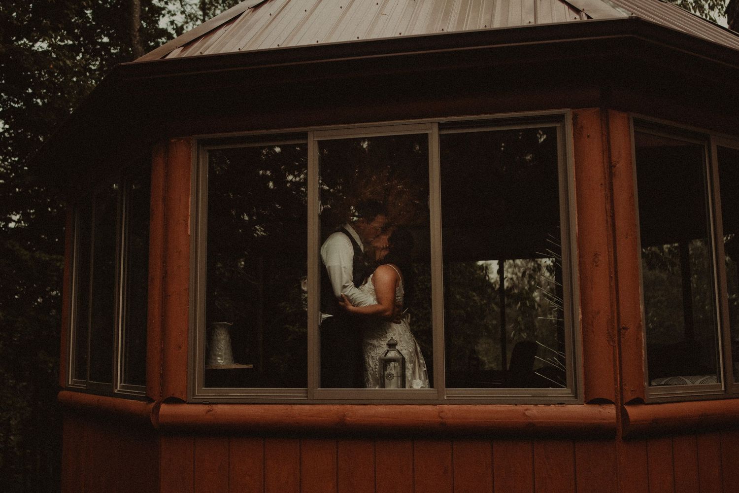 A wooden gazebo with large windows and a metal roof sits nestled among trees in a wooded setting.