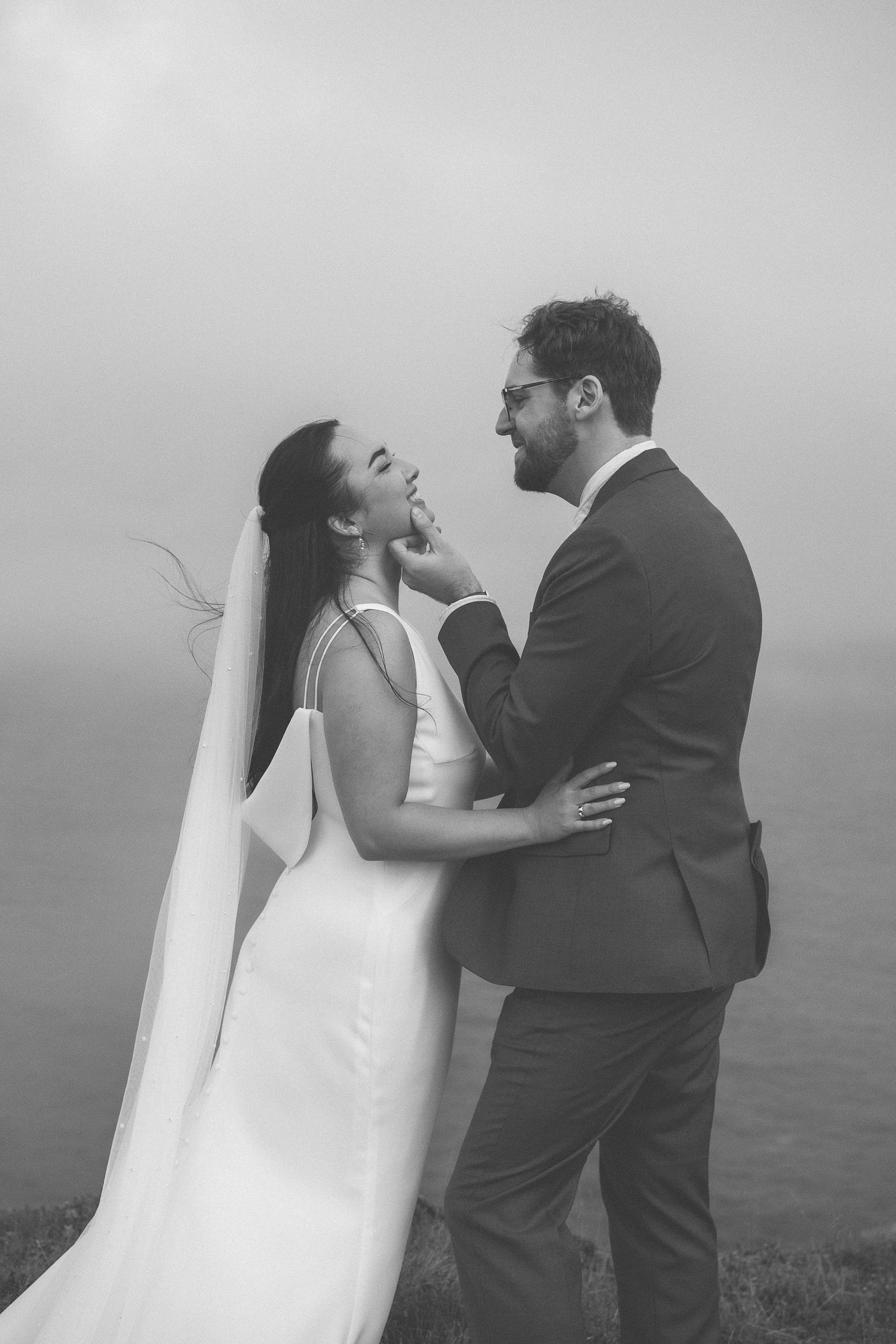 A couple stands on a coastal cliff edge in black and white, with the bride's white dress flowing in the wind against the ocean.