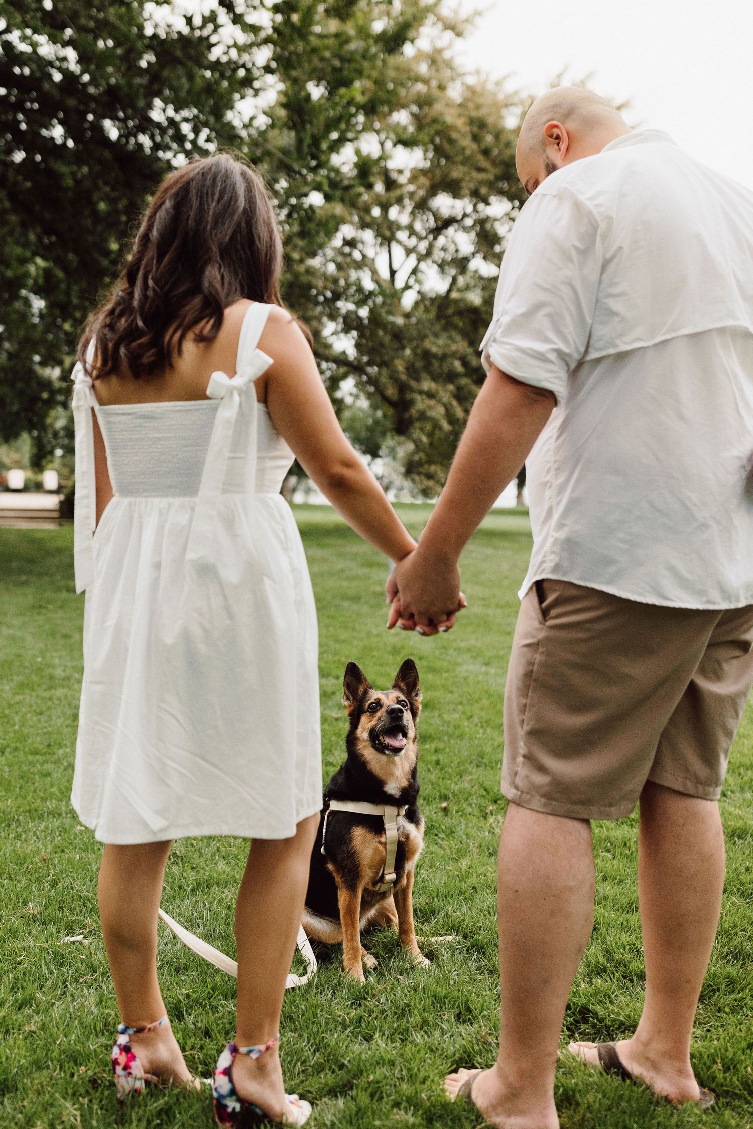 A couple in white and khaki clothing stands on grass with their German Shepherd dog.