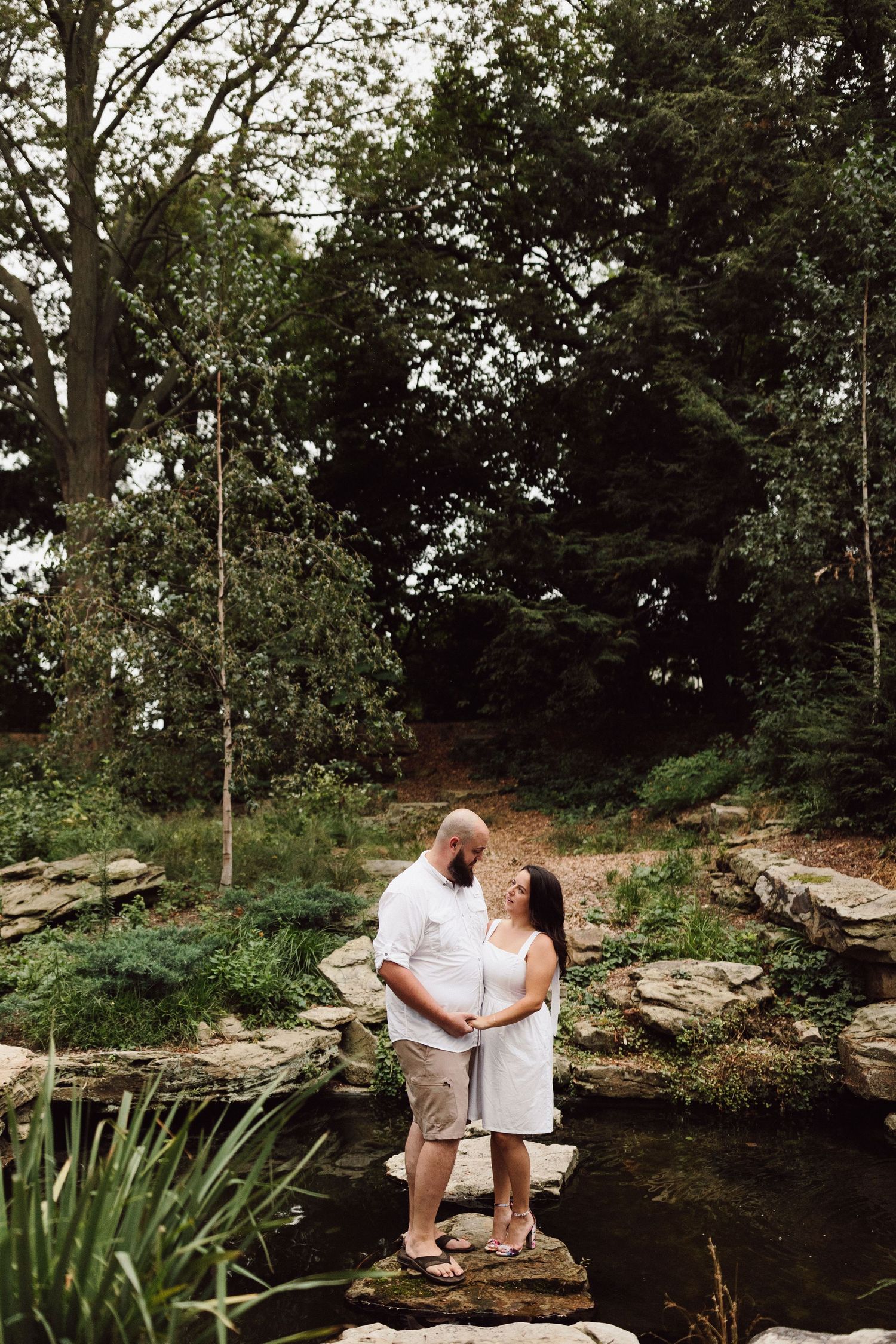 A couple sits together on rocky outcrops near a peaceful pond with trees in the background.