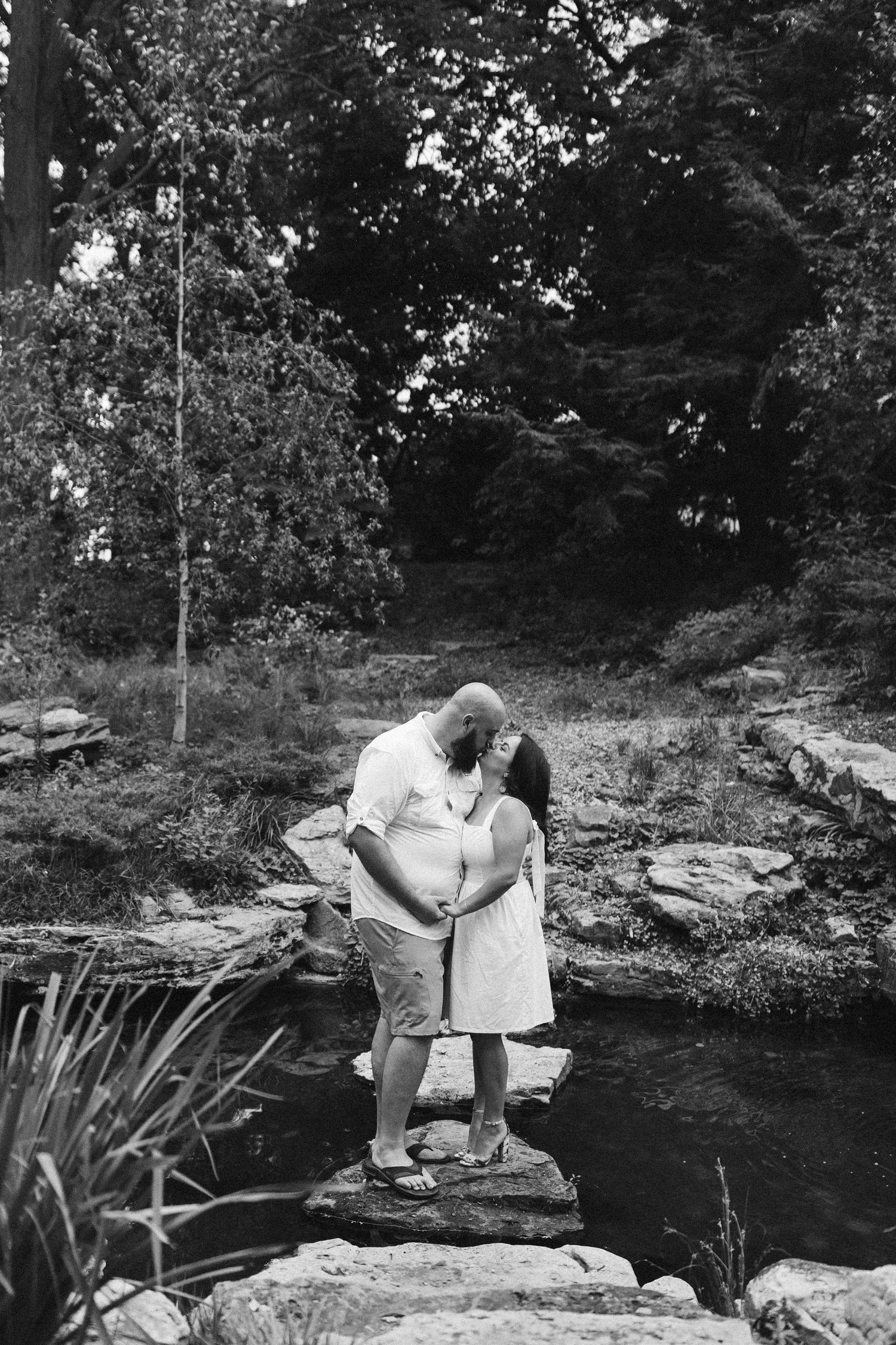 A black and white photo of a couple sitting on rocks by a peaceful pond.