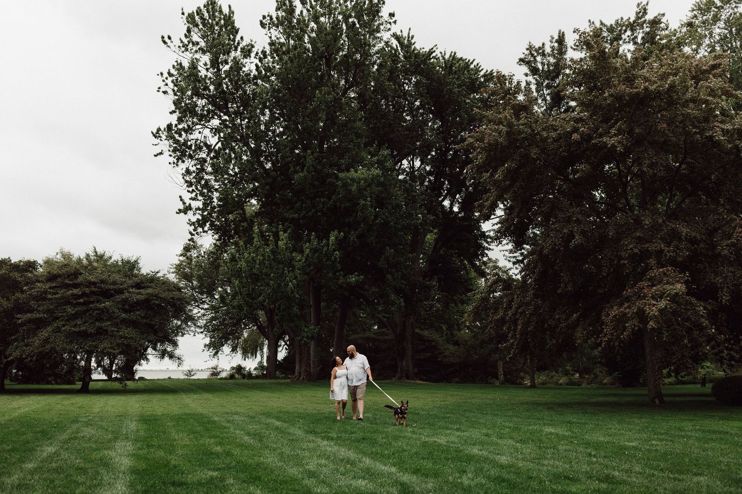 A couple walks with their dog across a vast green lawn surrounded by mature trees.