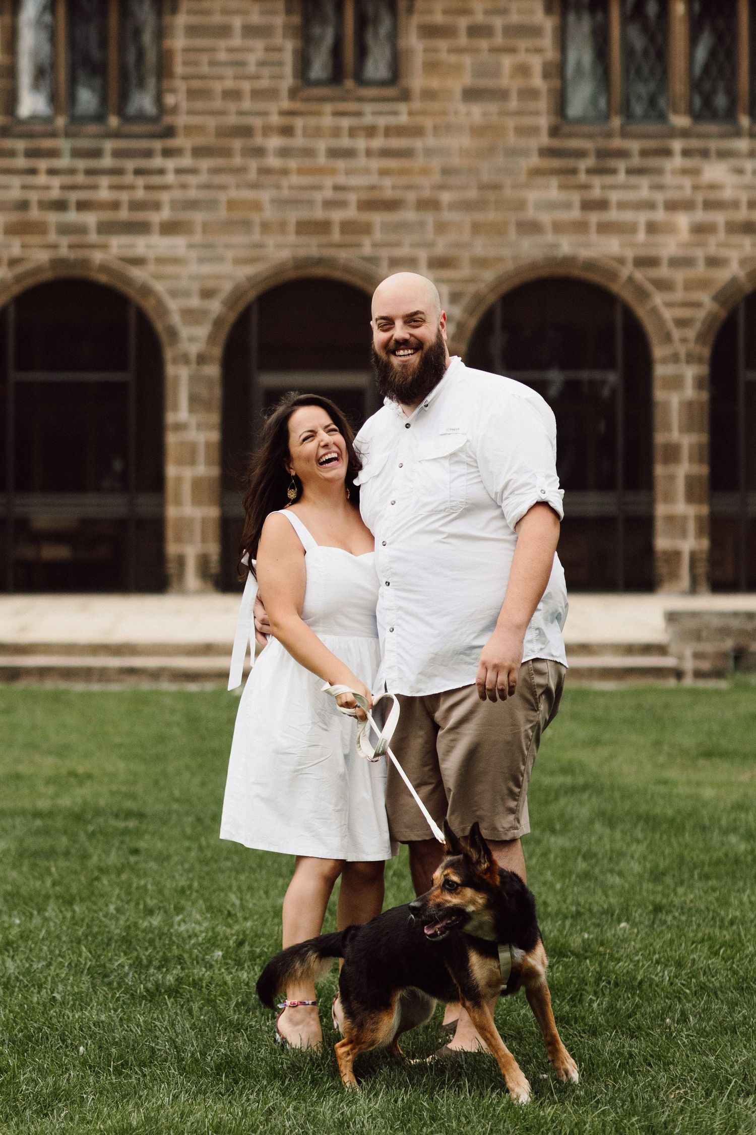 A couple stands with their small dog on a lawn in front of a Gothic stone building.