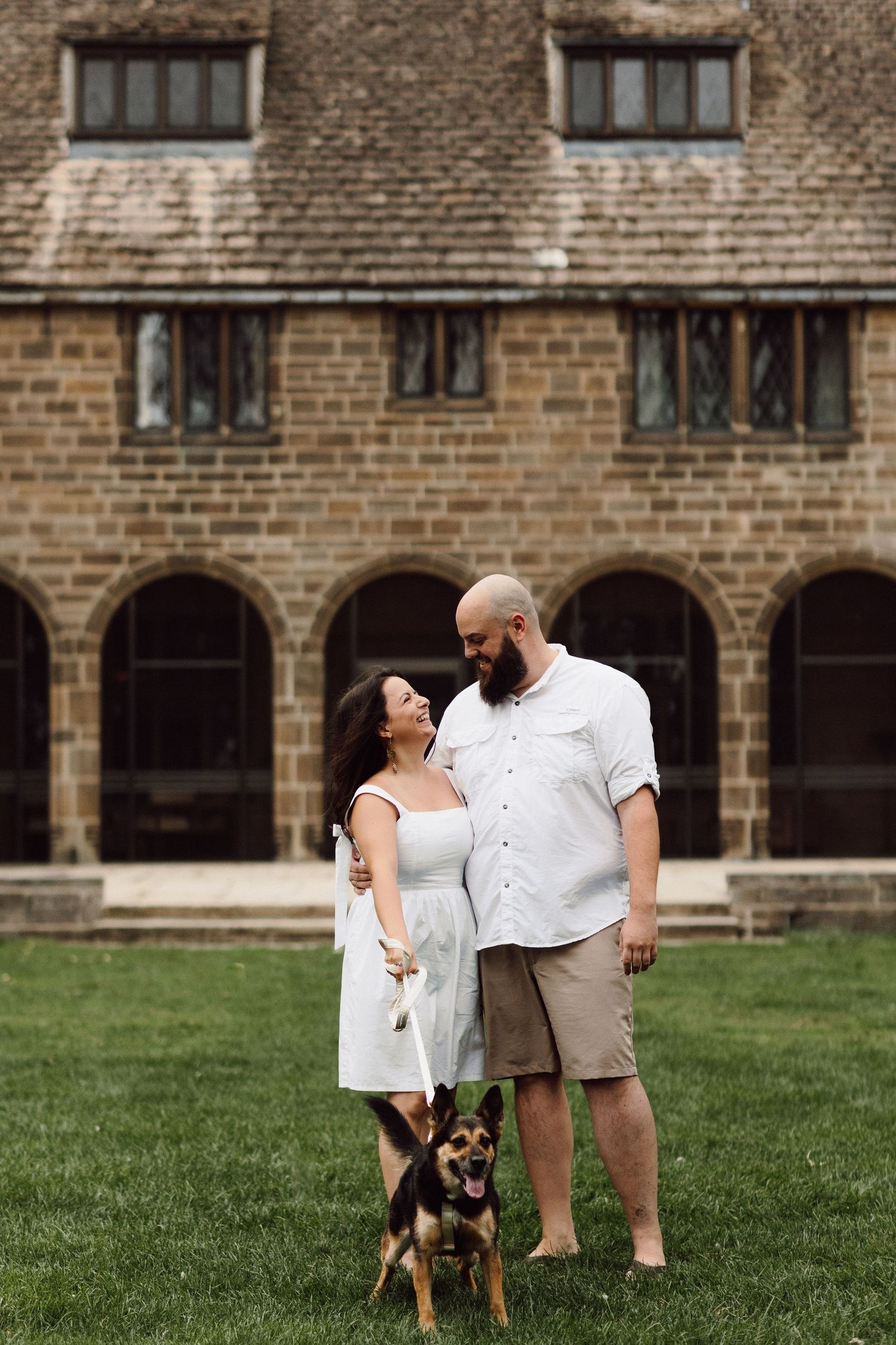 A couple poses with their dog against the backdrop of a historic stone building with arched windows.