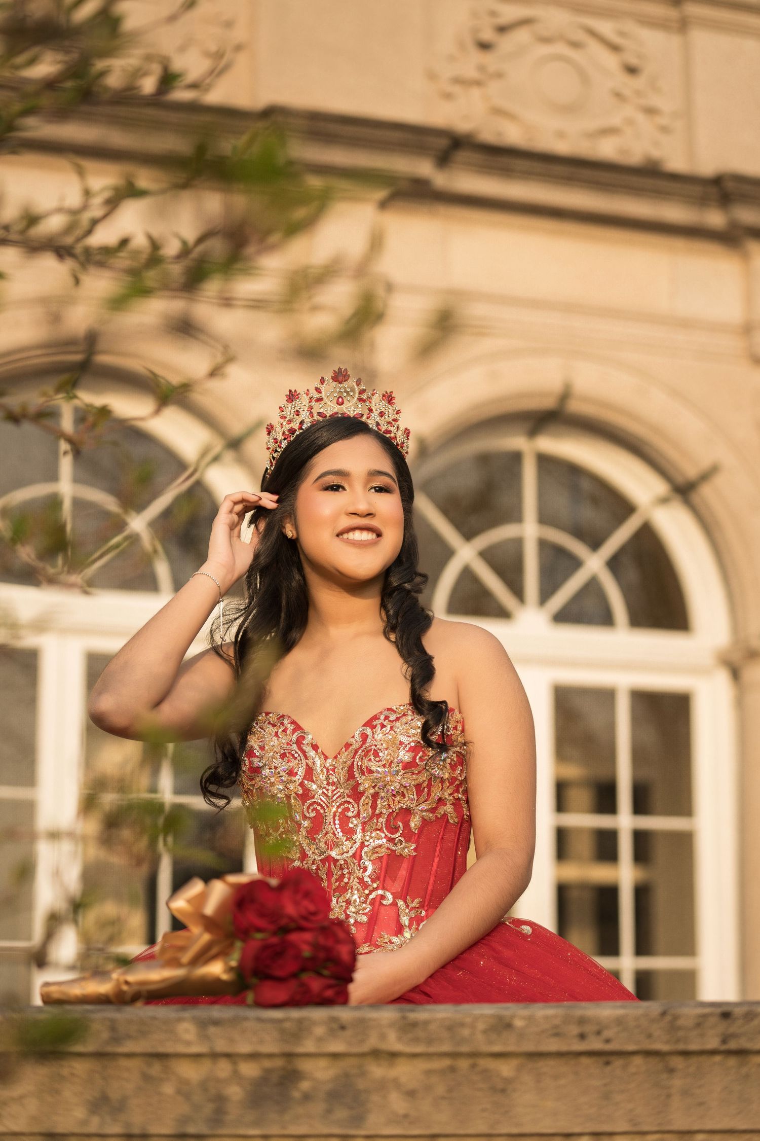 A person in a red embellished gown poses elegantly on a balcony with arched windows in the background.