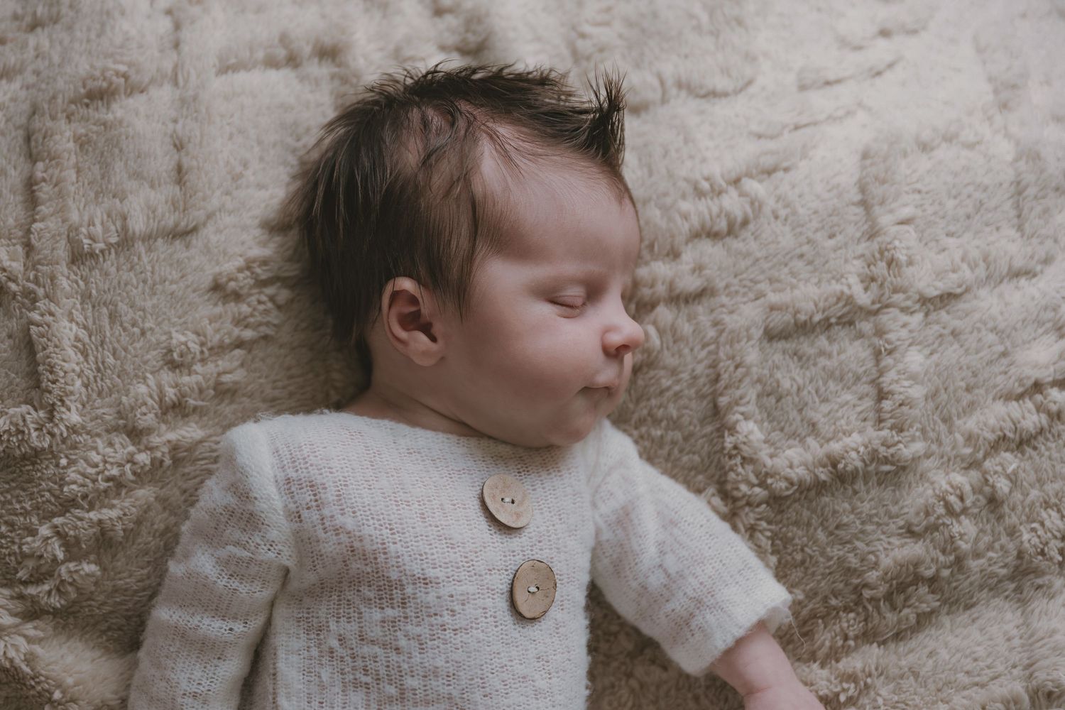 A sleeping baby wearing a cozy cream-colored cardigan sweater with wooden buttons lies against a textured beige wall.
