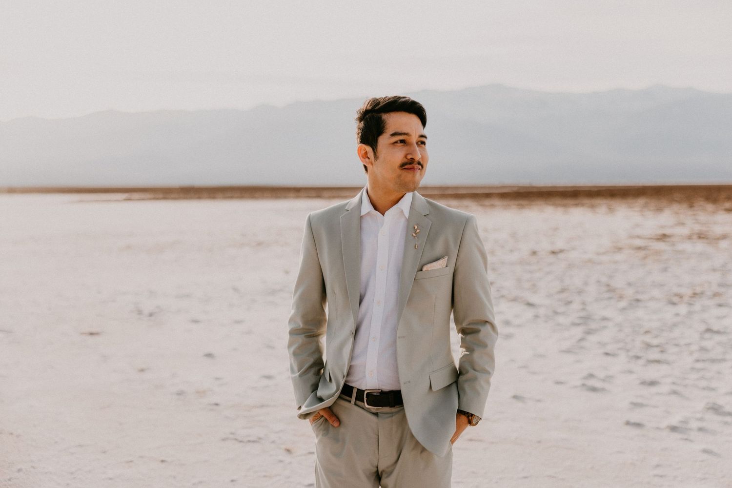 A groom poses in a light colored suit against a desert salt flat and mountain backdrop.