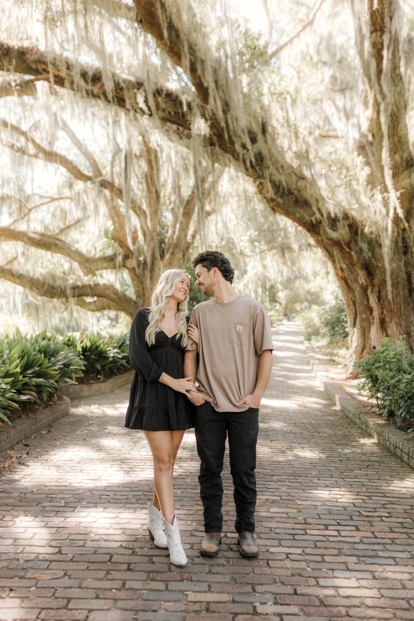 Couple smiling at each other on a brick path lined with moss-draped oak trees in a lush garden setting in Tallahassee Florida.