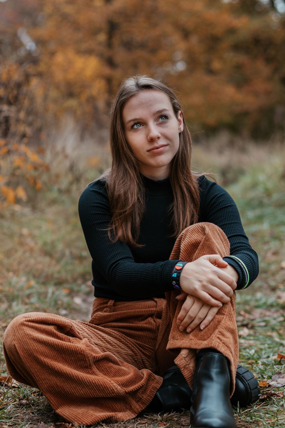Person in dark sweater and brown pants poses on autumn ground.