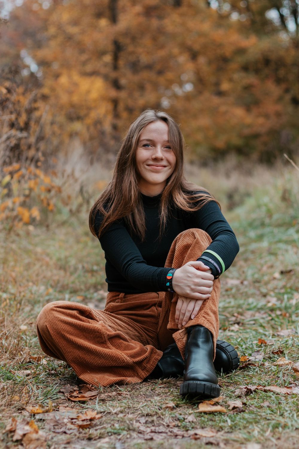Person in black sweater and brown pants poses on autumn ground.