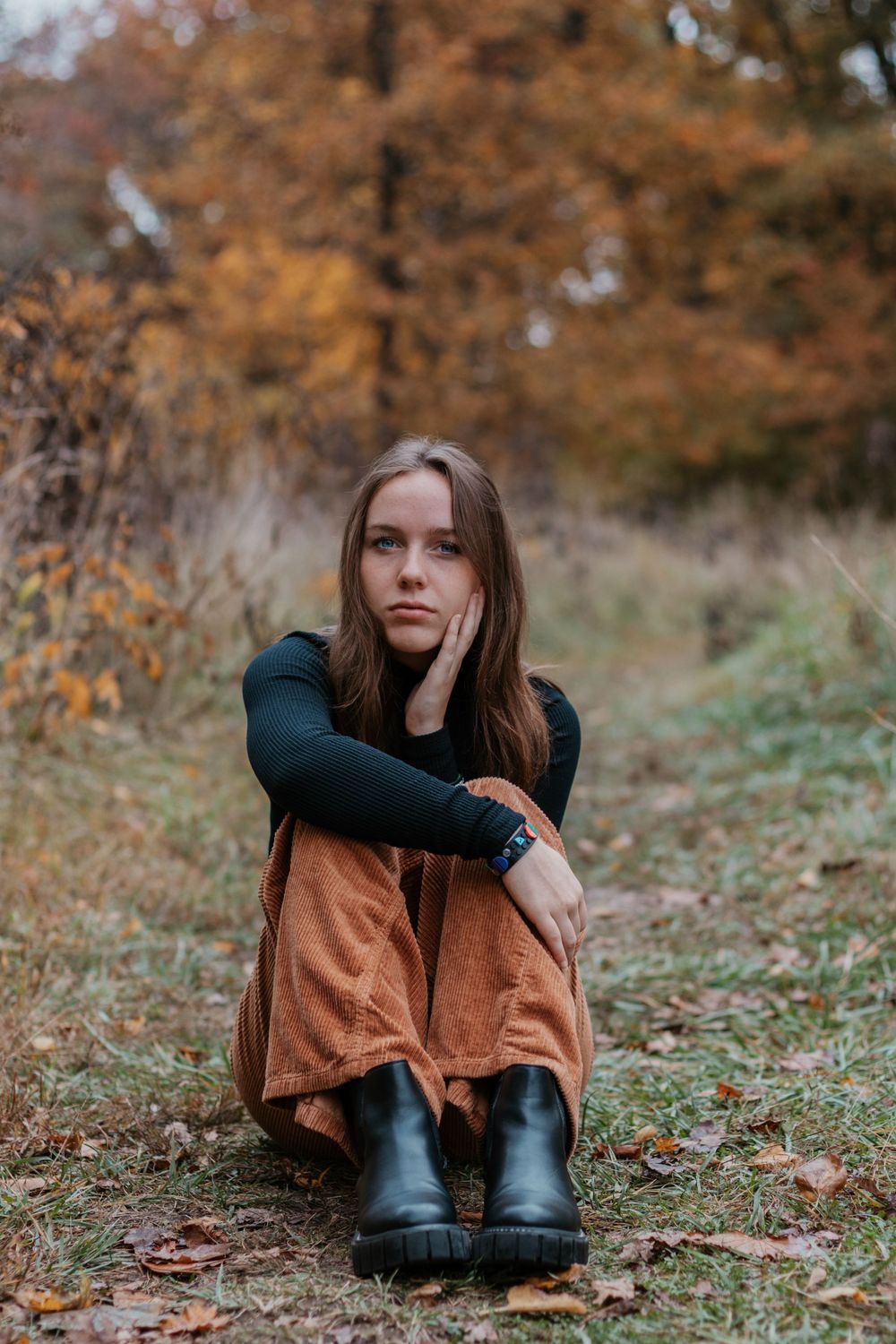 Person in dark clothing and boots sits on ground surrounded by fall foliage.