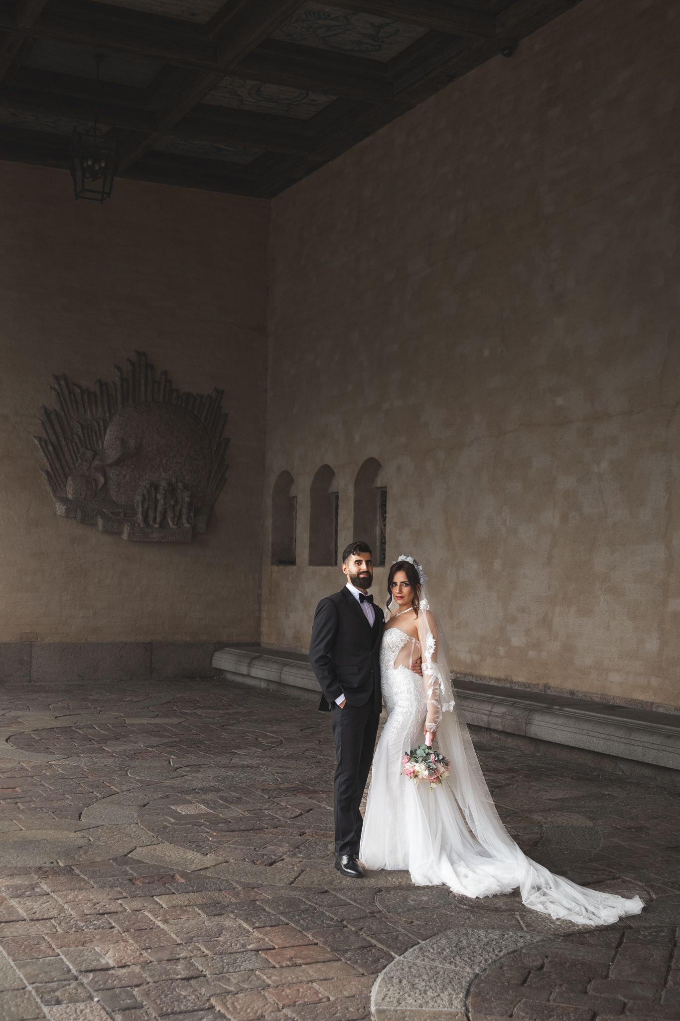 A wedding couple poses in a dramatic stone corridor with arched ceilings and patterned tile floors.