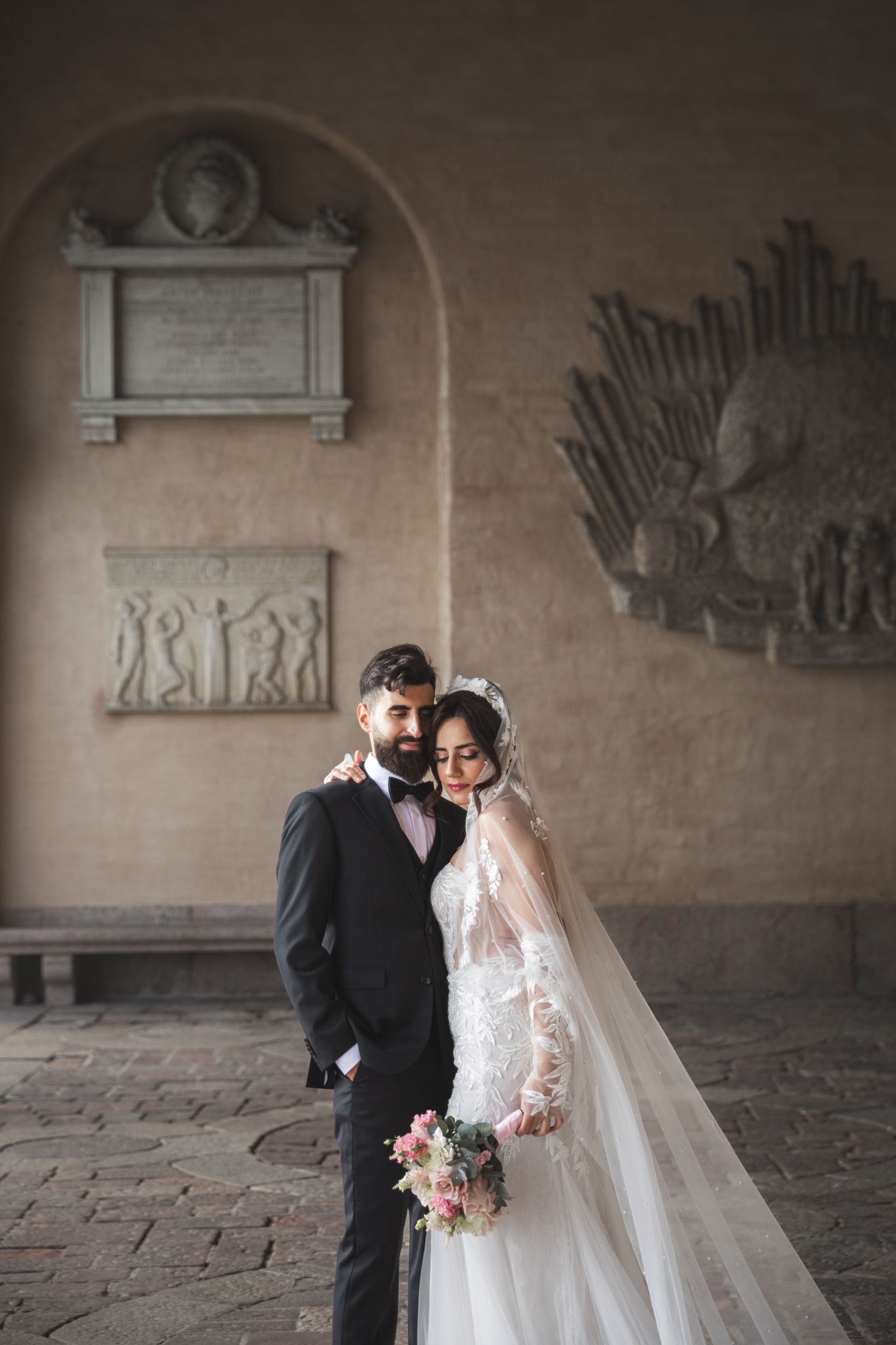Elegant wedding couple poses against stone wall in historic corridor with decorative architectural details.
