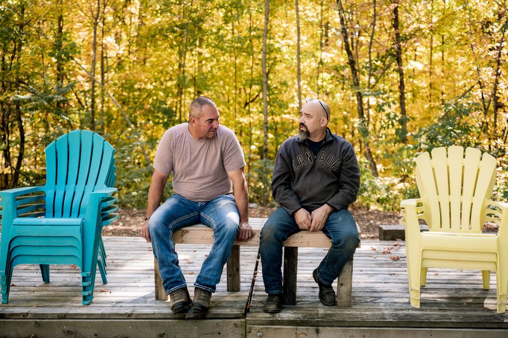 People sitting on colorful Adirondack chairs on a wooden deck surrounded by autumn foliage.