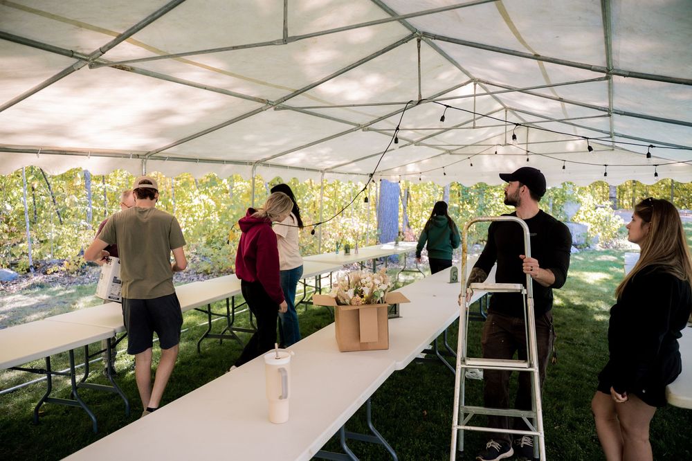 People setting up tables and decorations under a white outdoor event tent with natural lighting and greenery in background.