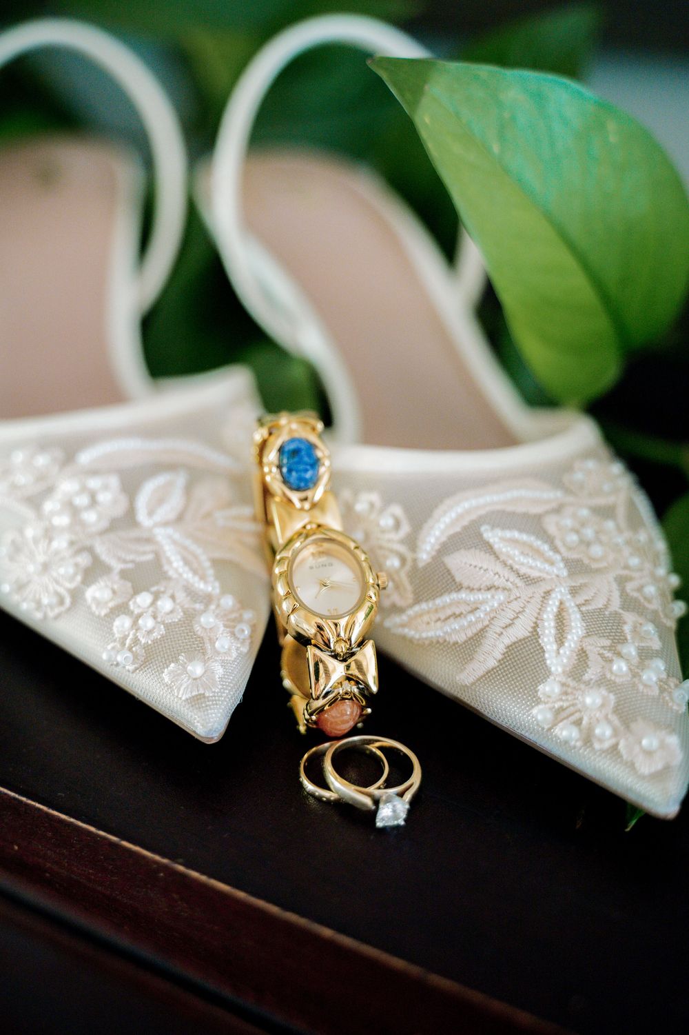 Elegant bridal shoes with lace embroidery displayed with wedding rings and jewelry on dark surface.