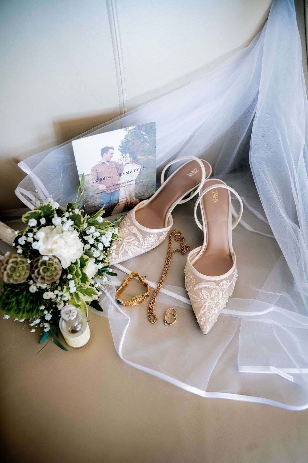 Elegant bridal accessories including white lace shoes, floral bouquet, and delicate veil arranged on soft fabric.