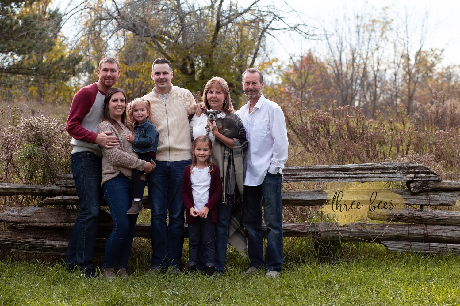 A group gathers for outdoor autumn family portraits by rustic wooden fences surrounded by fall foliage.