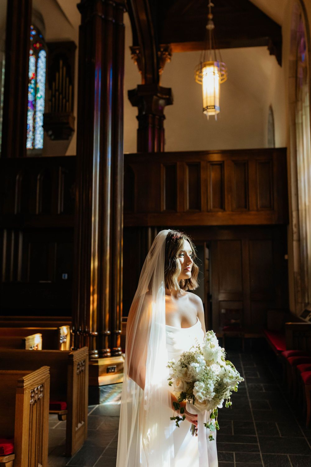 A bride in a white wedding dress holds a bouquet of white flowers inside a historic church with wooden paneling and stained glass.