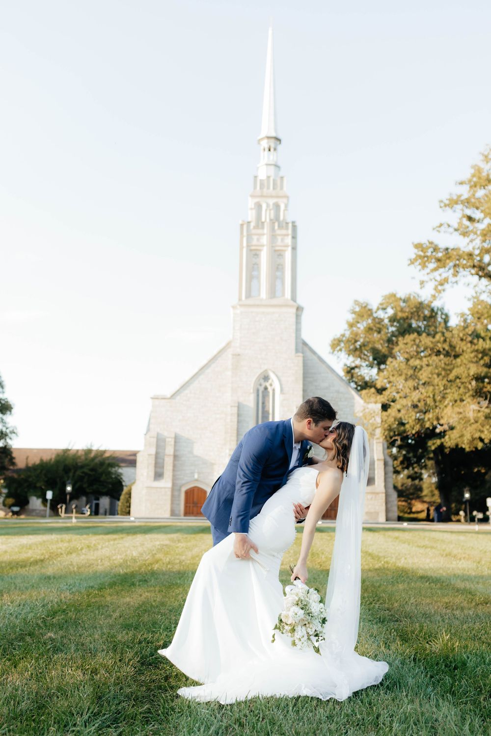 A couple shares an intimate moment on a grassy hill with a white church steeple visible in the background.