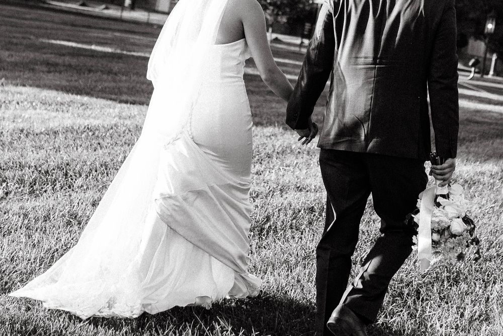 Black and white photo of a bride in flowing white dress and groom in dark suit walking on grass during a wedding ceremony.