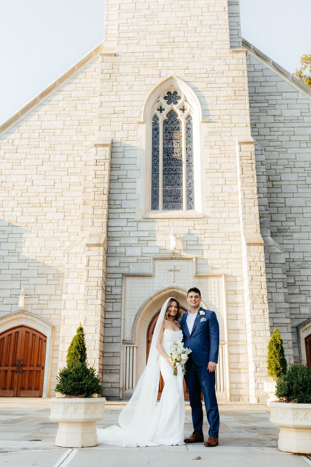 A wedding couple poses in front of a beautiful white stone church with Gothic windows and wooden doors.