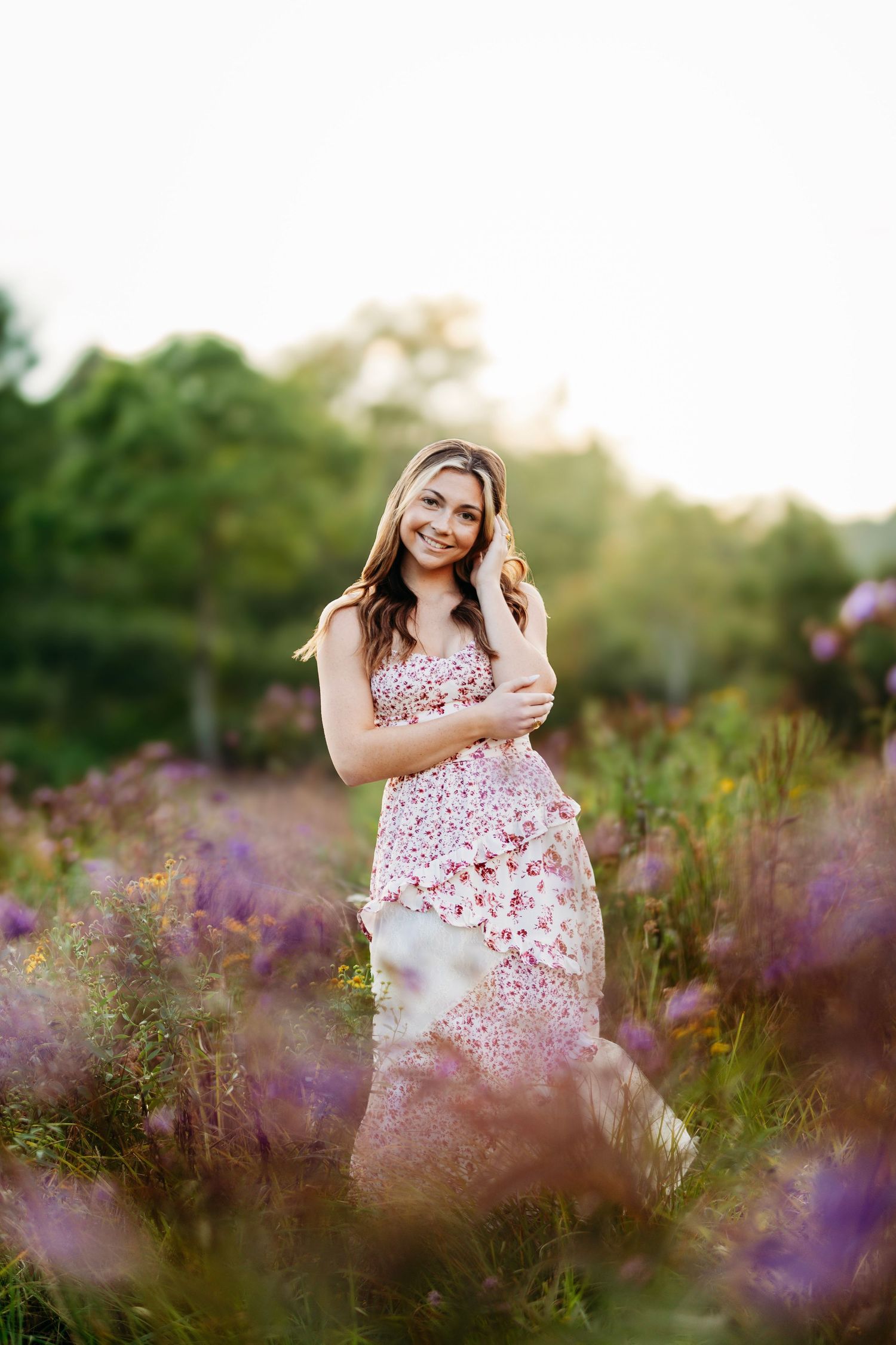 Young woman in floral dress standing in purple wildflower field during golden hour sunset.