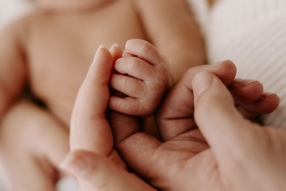 Close up photo of gentle hands holding a tiny newborn baby's fingers against a soft white background.