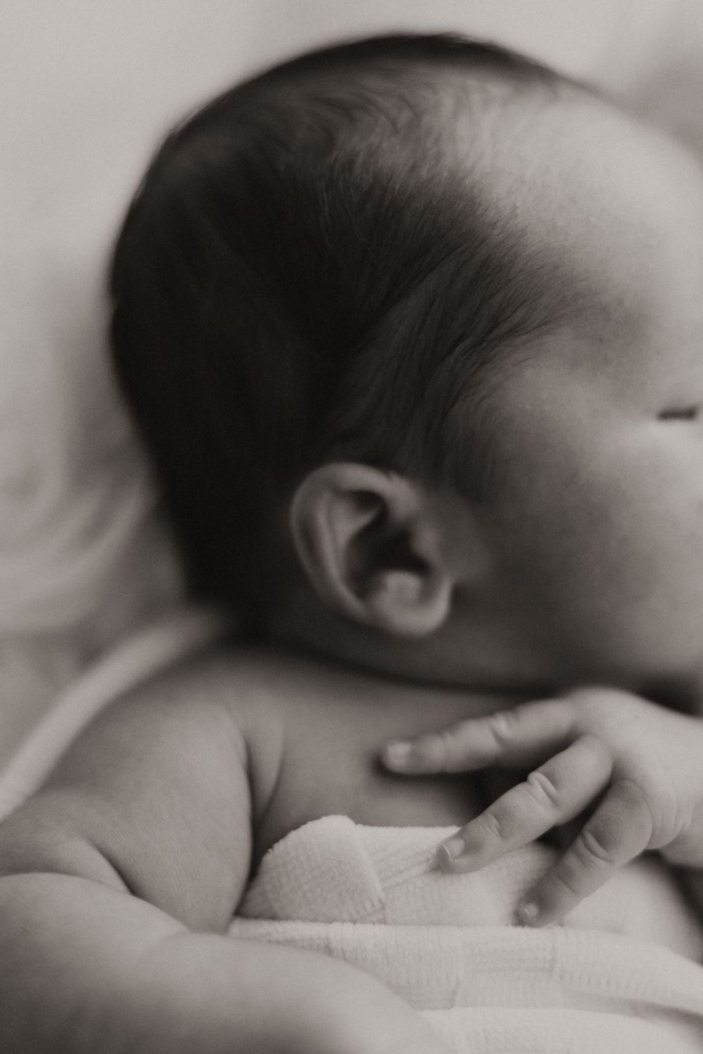 Close-up black and white photo of a sleeping baby's head and tiny hand resting on soft fabric.