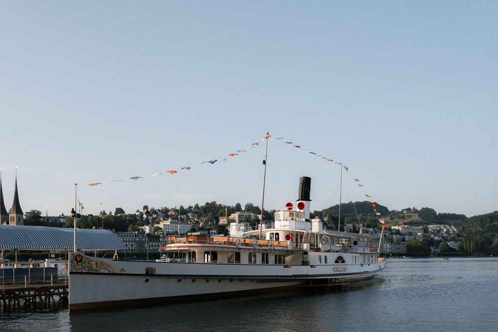 A panoramic view of boats docked at a waterfront marina with mountains and buildings in the background. Luzern Schifffahrtsgesellschaft