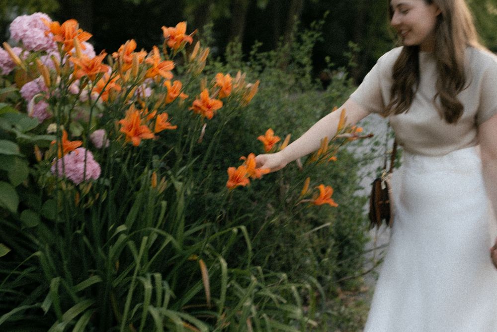 A person in a white dress poses among vibrant orange lilies in a lush garden setting.