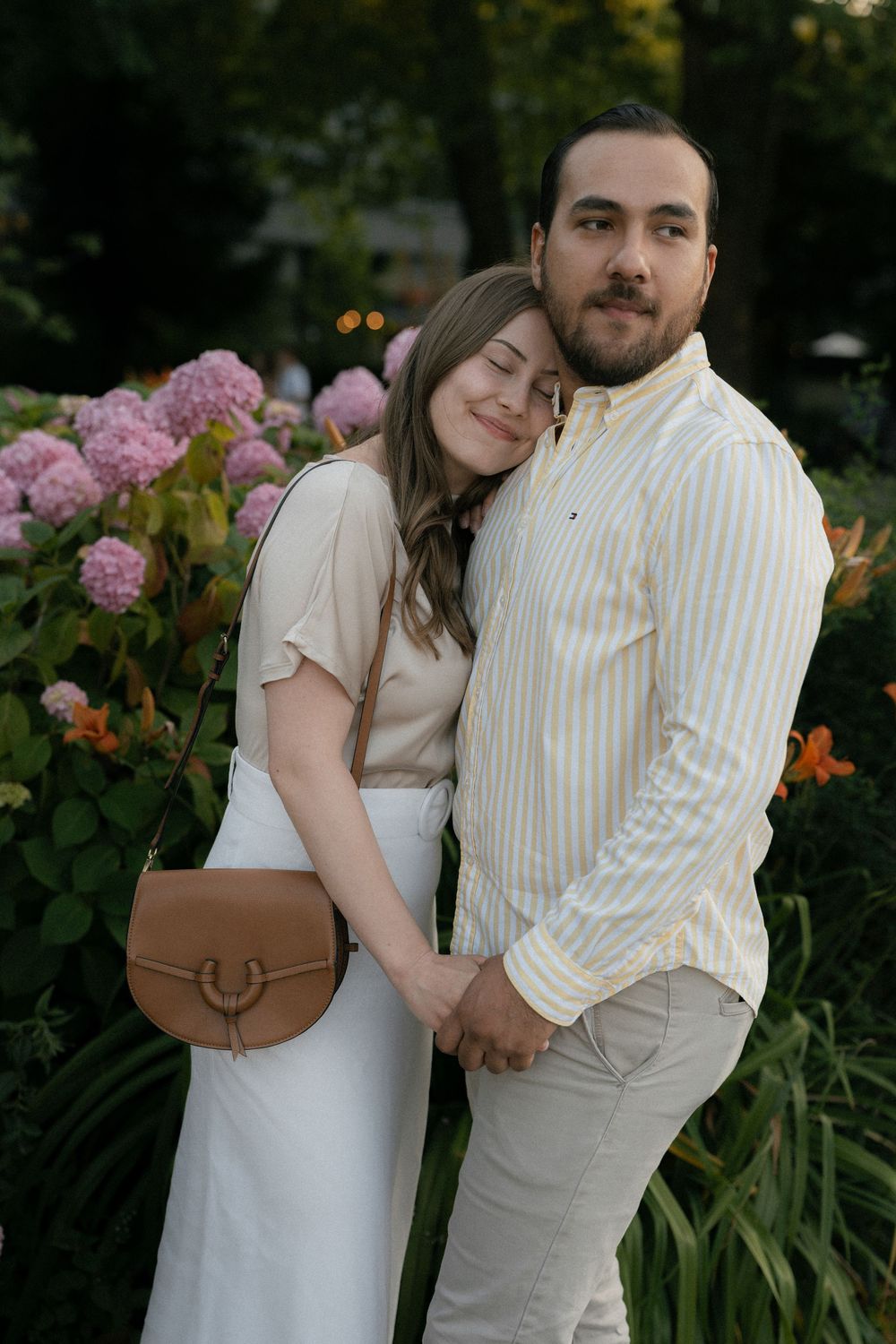 A romantic couple poses together in a garden with pink hydrangeas and a brown leather purse.