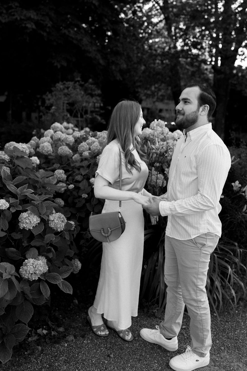 Couple in white outfits dancing near blooming hydrangea bushes in a garden.