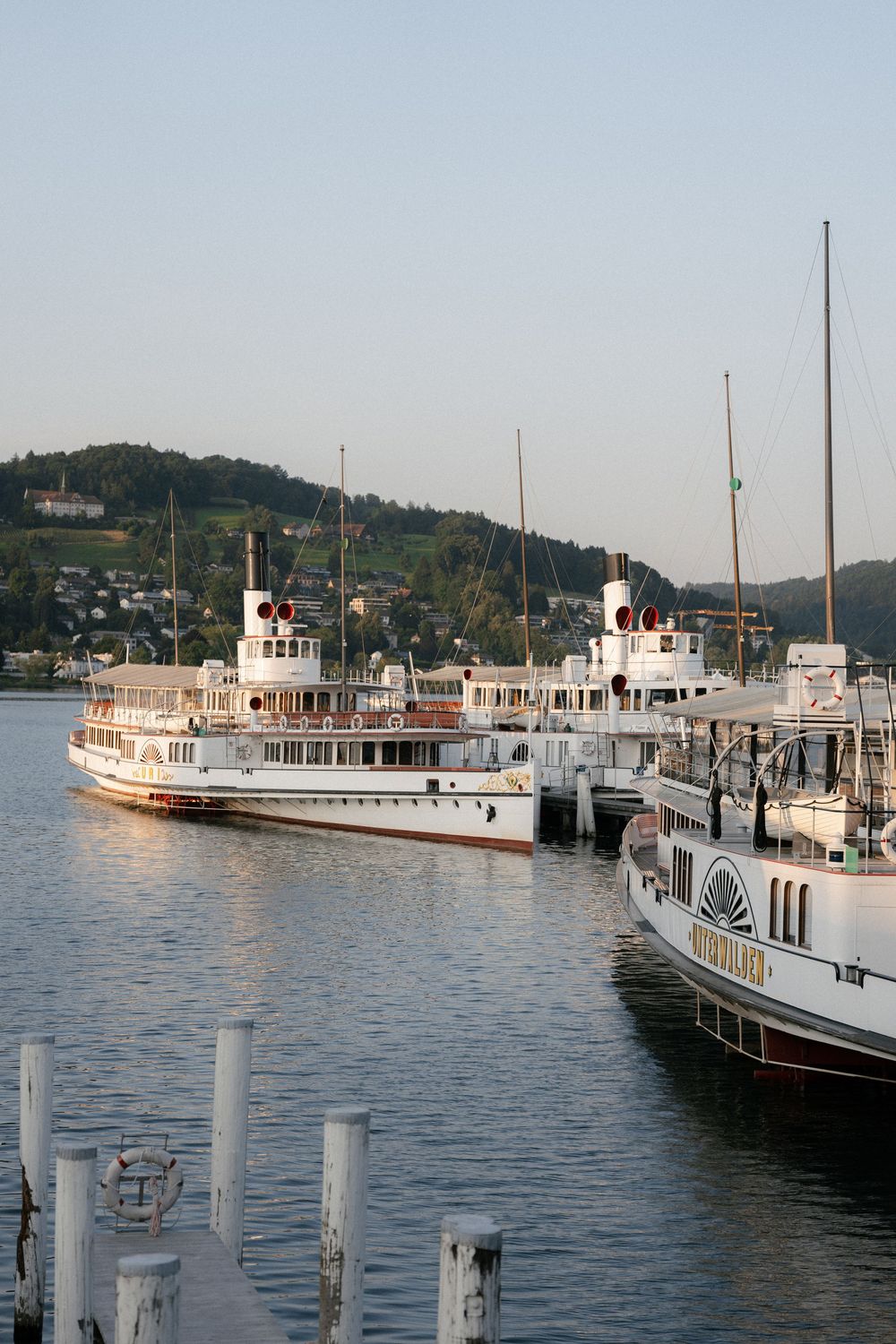 Vintage white passenger boats docked at a marina with mountains in the background during sunset. Luzern